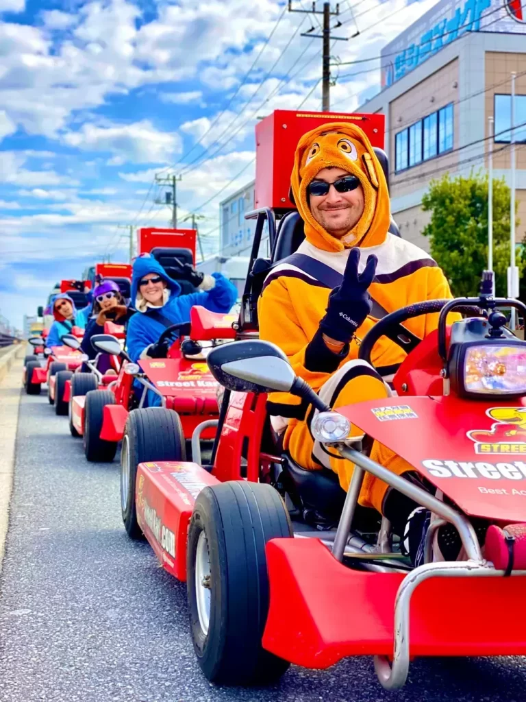 IMG_2817 - StreetKart Person in an orange animal-costume hoodie driving a red go-kart, flashing a peace sign in a parade line down a city street.
