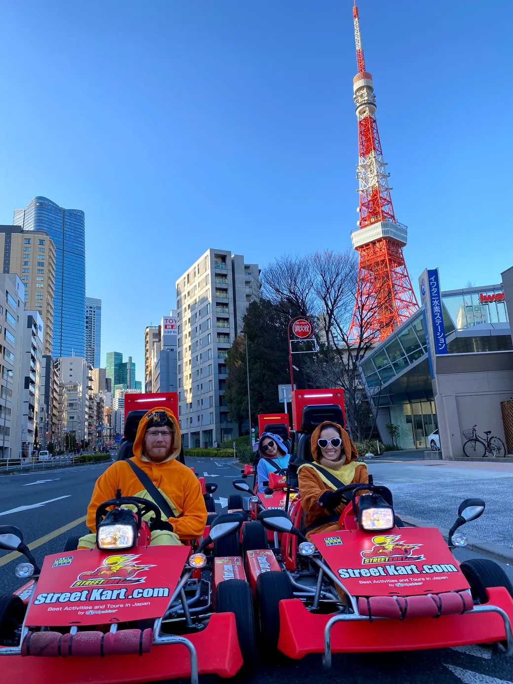 Group of people in orange costumes riding red go-karts on a city street with Tokyo Tower in the background.
