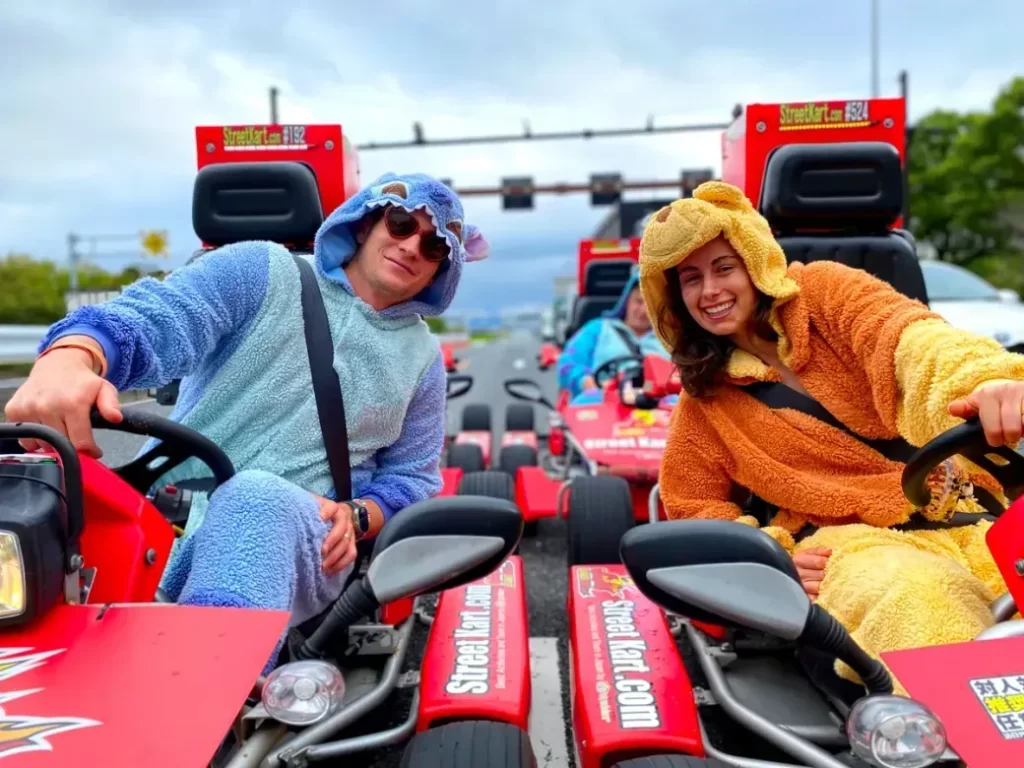 Two people in blue and orange animal onesies drive red go-karts on a road track, smiling at the camera.